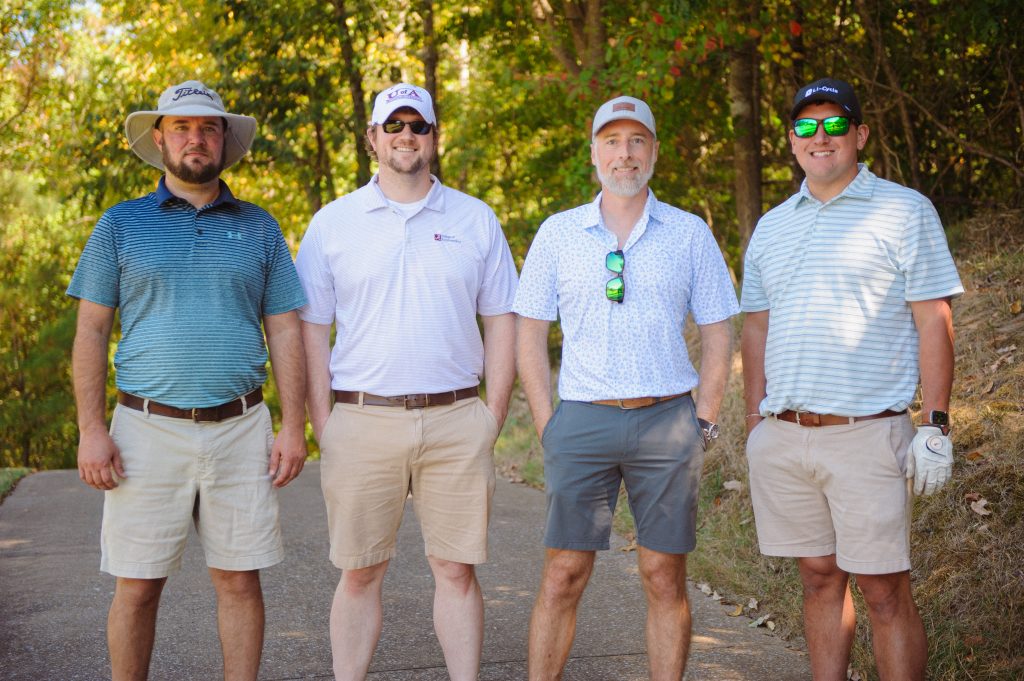 4 guys, 2 in stripes, 1 in patterned shirt, stand on a road like area on a golf course