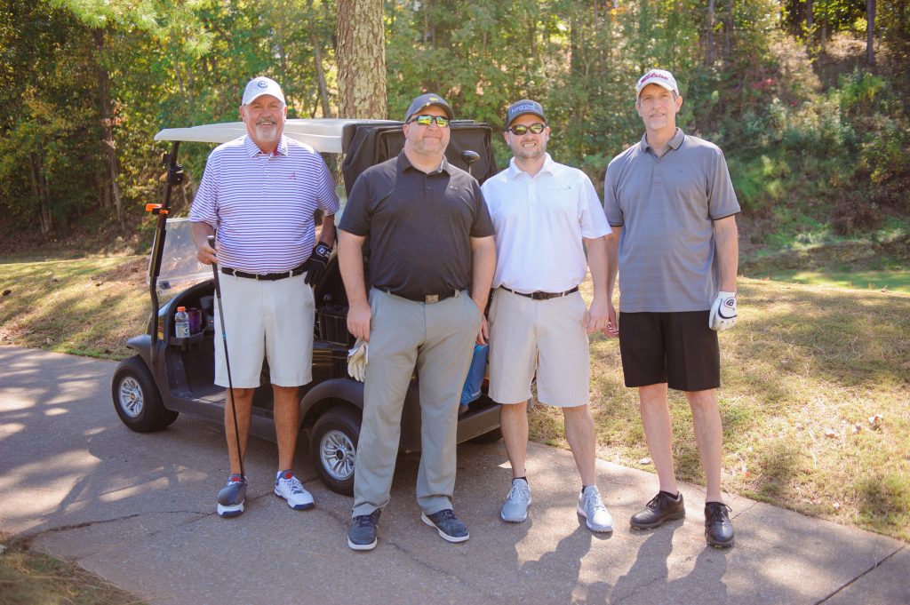 4 guys in various shades of grey to white shirts standing on a golf course