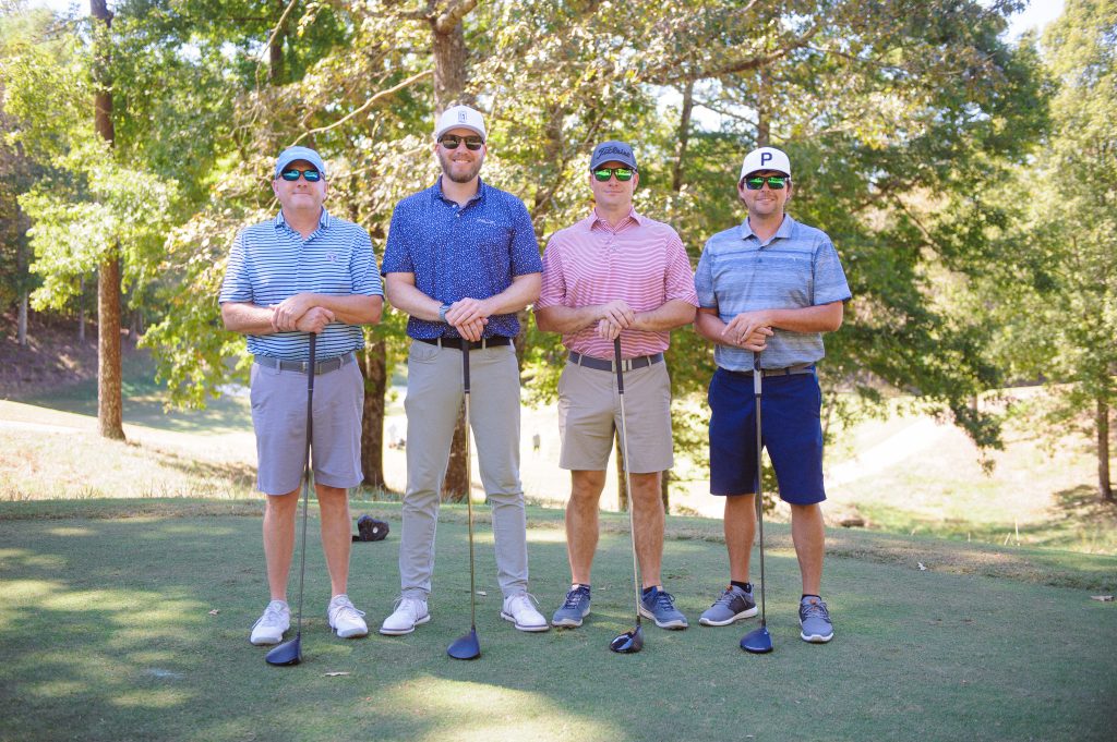 4 guys, 3 in blue shirts standing on a golf course