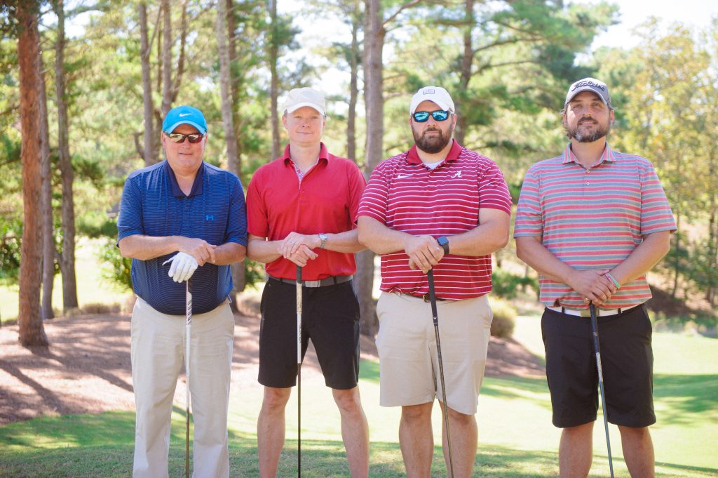 4 guys, 1 in blue shirt, 1 in red shirt, 1 in red and white stripes shirts standing on a golf course