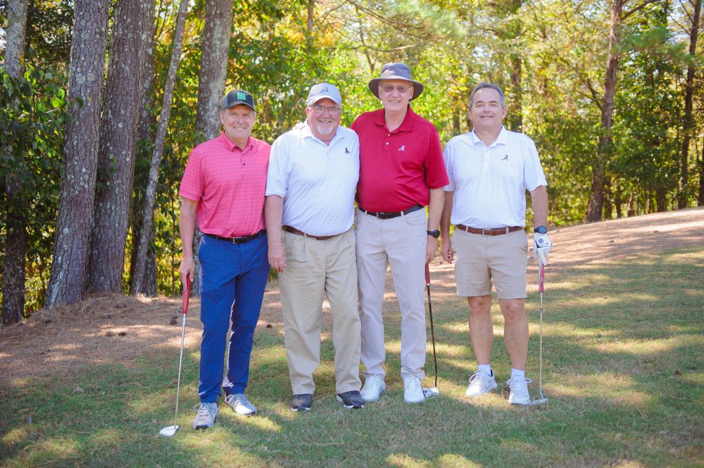 4 guys, 1 in pink shirt, 1 in red shirt, 2 in white shirts standing on a golf course