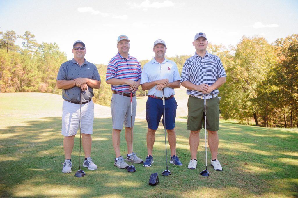 4 guys, 1 in striped shirt, standing on a golf course